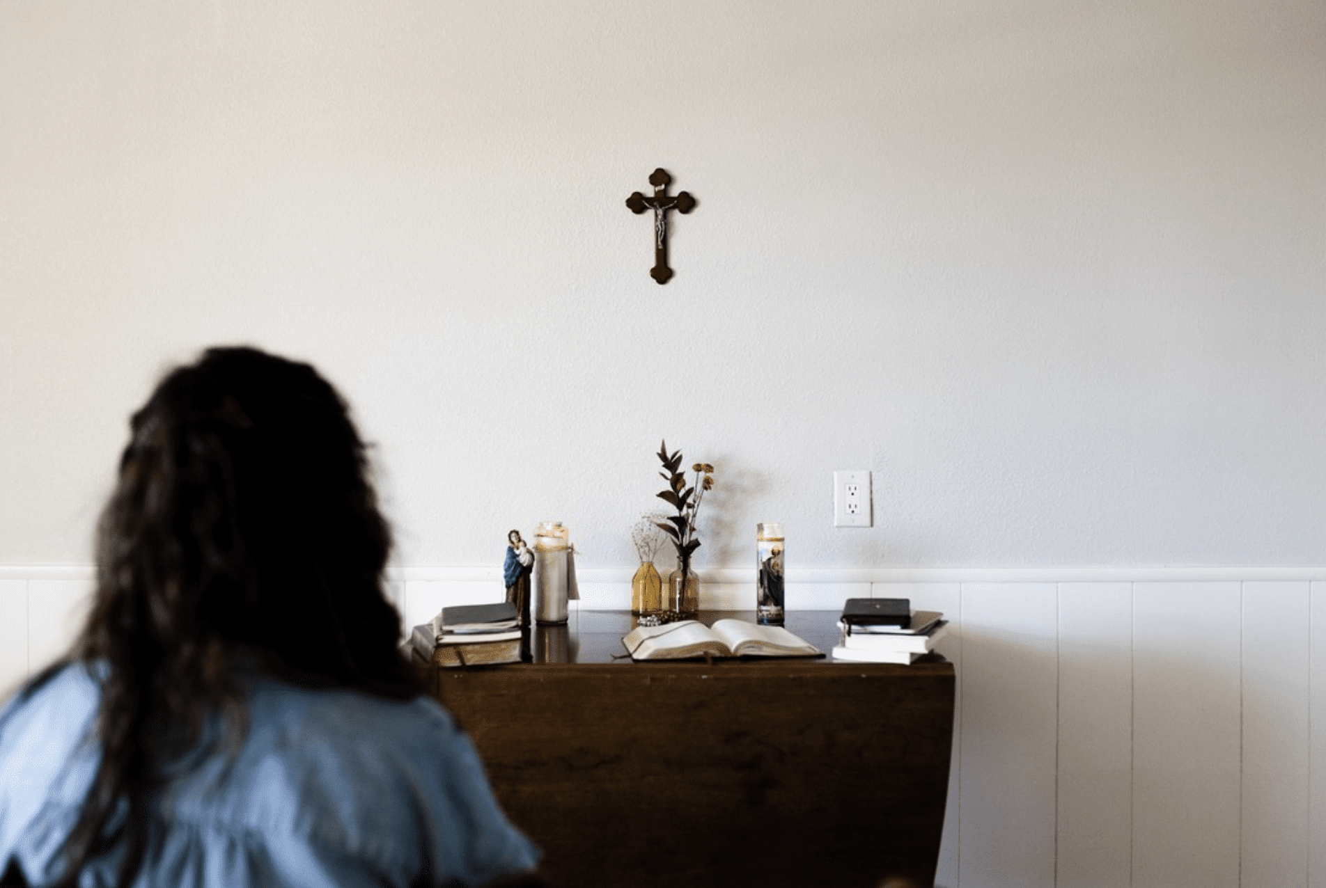 A girl prays before an altar with books and sacred objects in a home prayer space. 